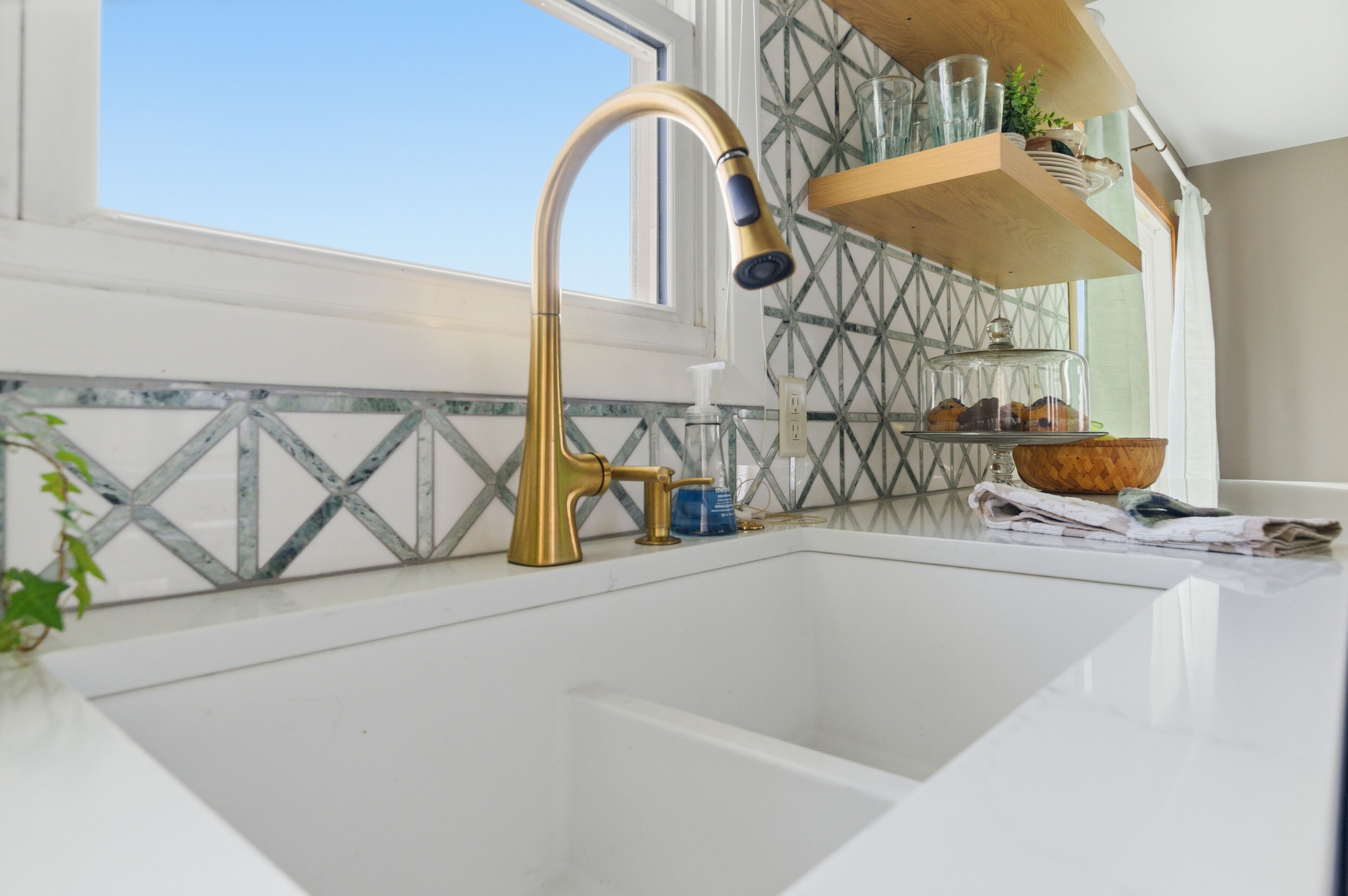 Eine Küche mit weißer Arbeitsplatte, einem goldenen Wasserhahn, einem Fenster und einem Regal mit Gläsern und einer Pflanze.