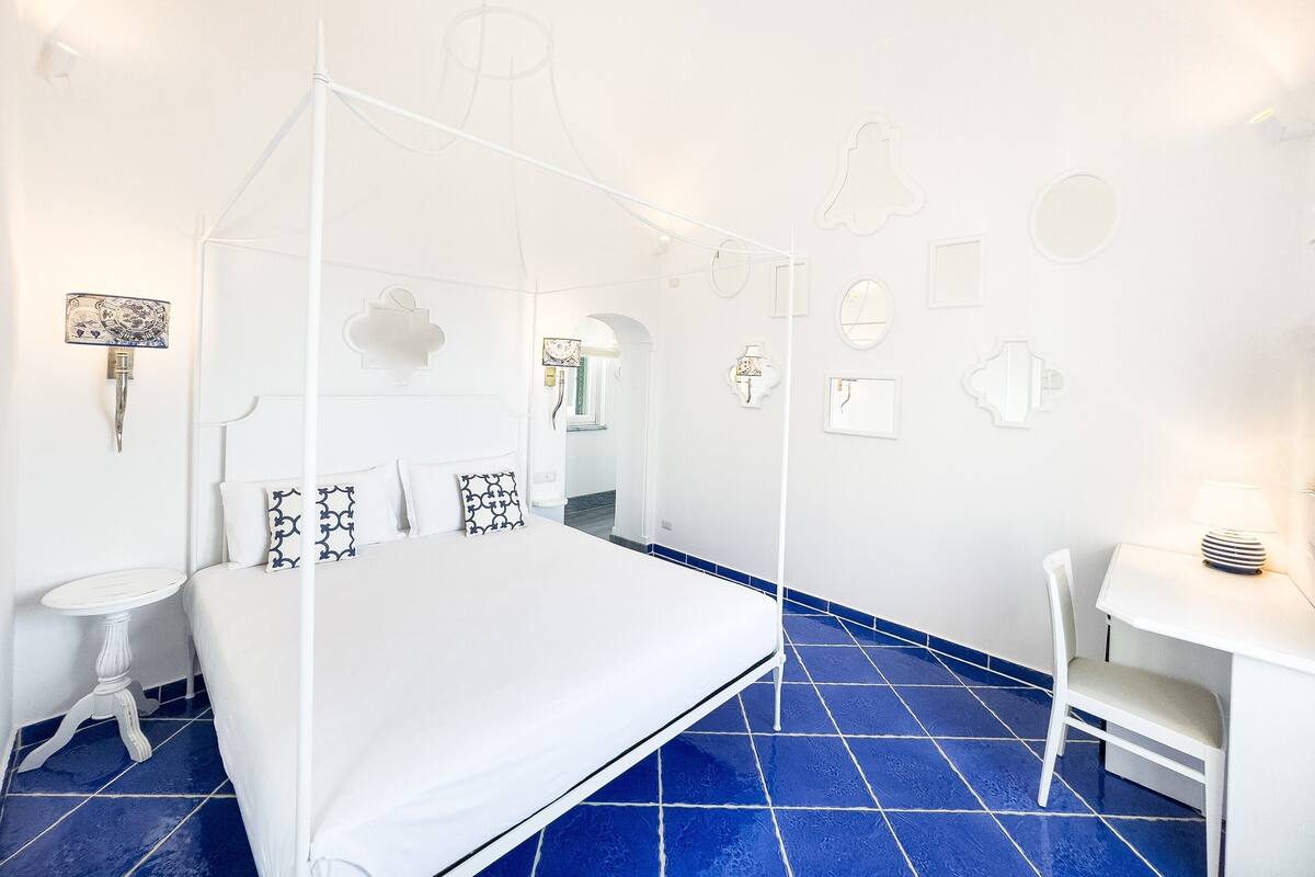 A bedroom with a four-poster bed, a small bedside table, a chair, and a desk with a lamp.