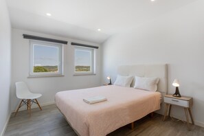 Un dormitorio con una cama, dos ventanas, una silla, una mesita de noche con una lámpara y vistas al exterior.