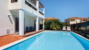 A swimming pool with a clear blue water, surrounded by a white building with a balcony and a red-tiled roof.
