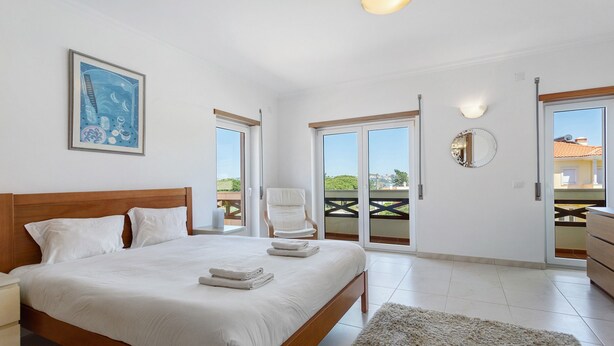 A bedroom with a bed, a nightstand, a chair, a large window, and a balcony.