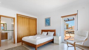 A bedroom with a wooden bed, a white chair, a nightstand, and a view of the outside.
