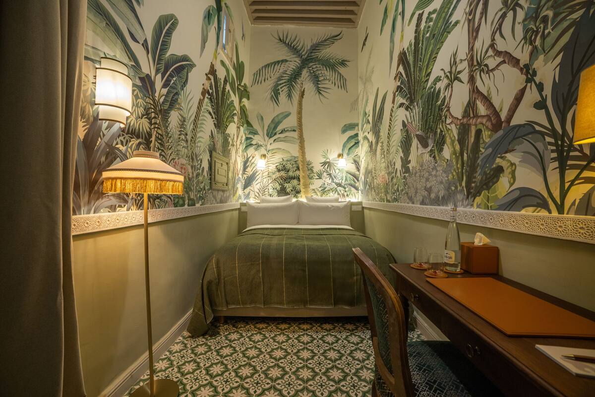 A small bedroom with a bed, a nightstand, a lamp, and a wall mural of tropical plants.