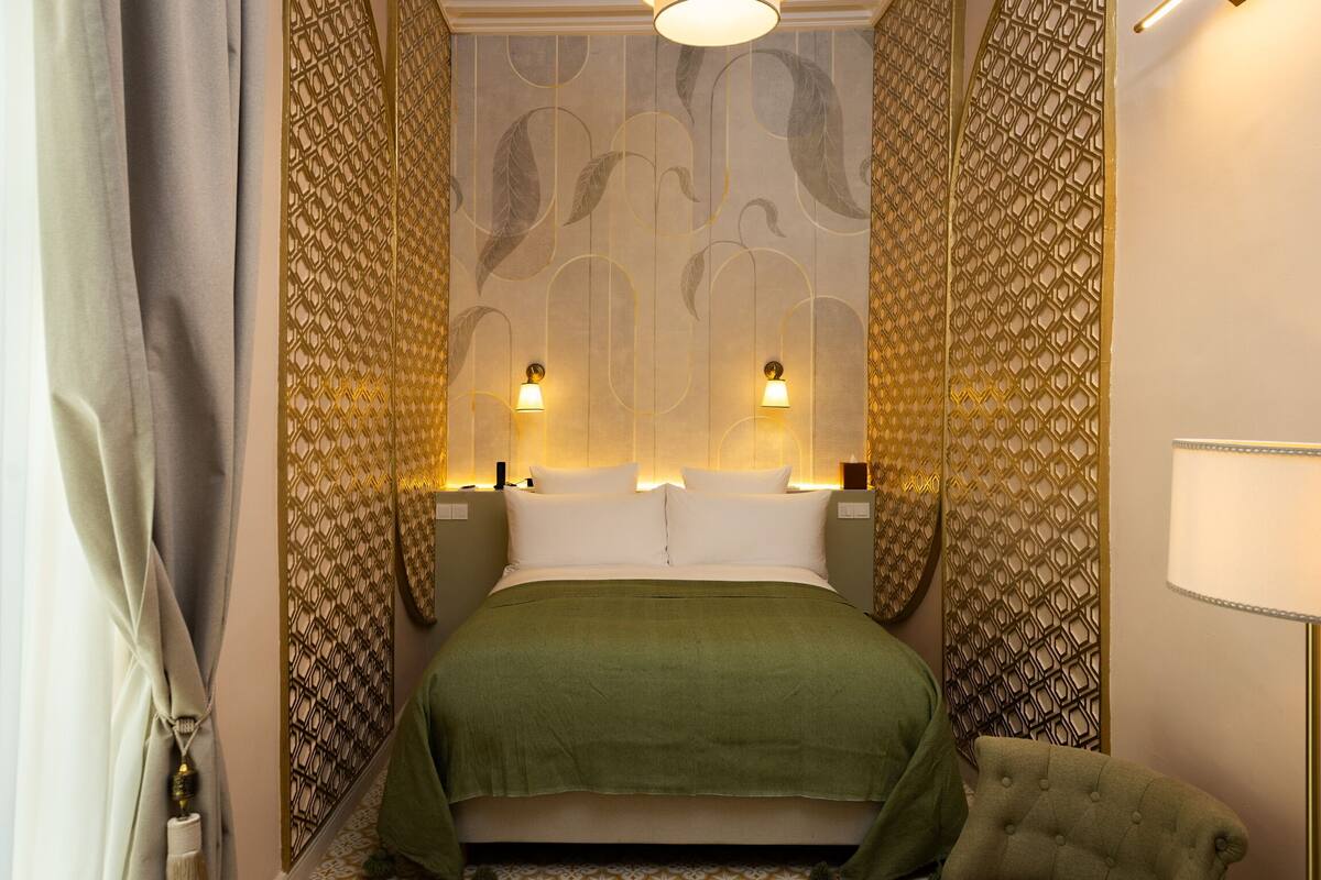 A bedroom with a bed, two wall-mounted lamps, a green bedspread, and patterned wallpaper.