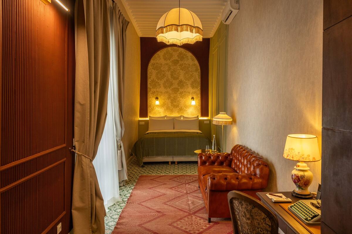 A hotel room with a bed, a leather sofa, a desk with a lamp, and a patterned rug.