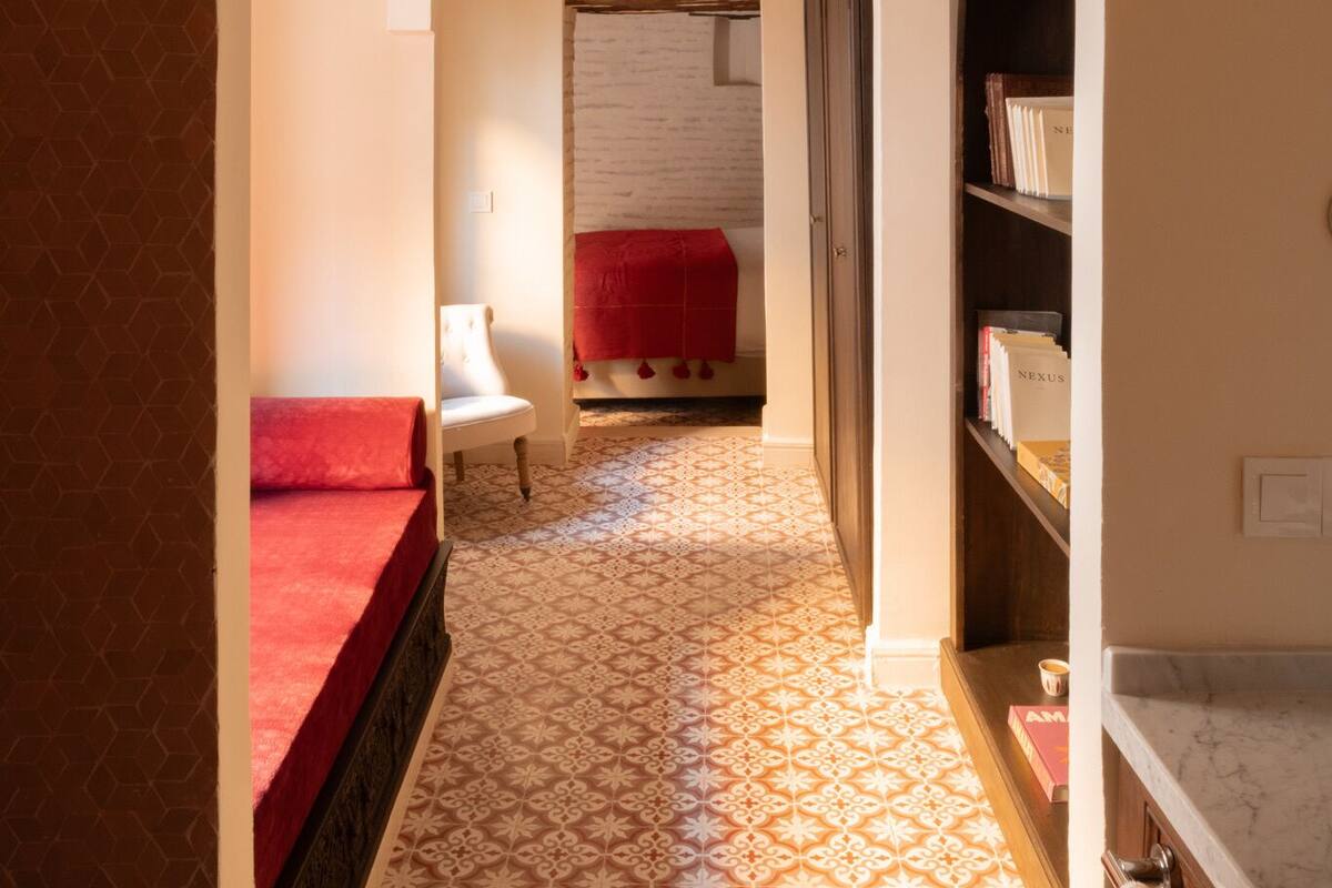 A hallway with a patterned floor, a red bed, a white chair, and a bookshelf.