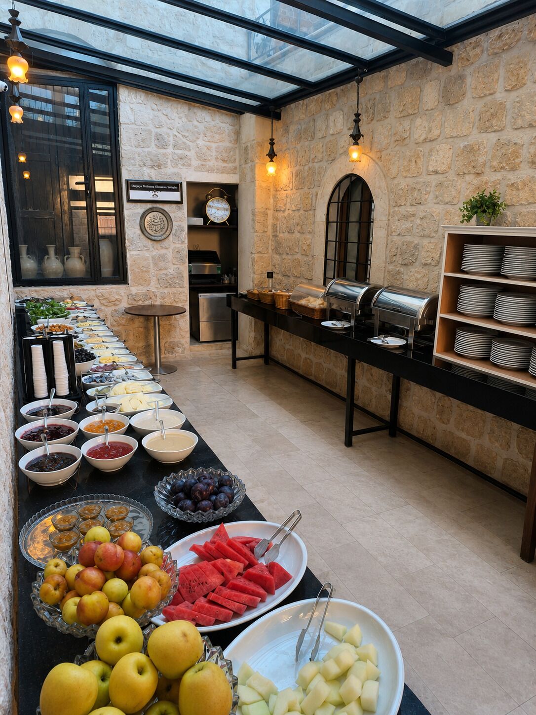 Montaje de bufé con diversas opciones de comida, un reloj y una estación de café en una sala de paredes de piedra con techo de vidrio.