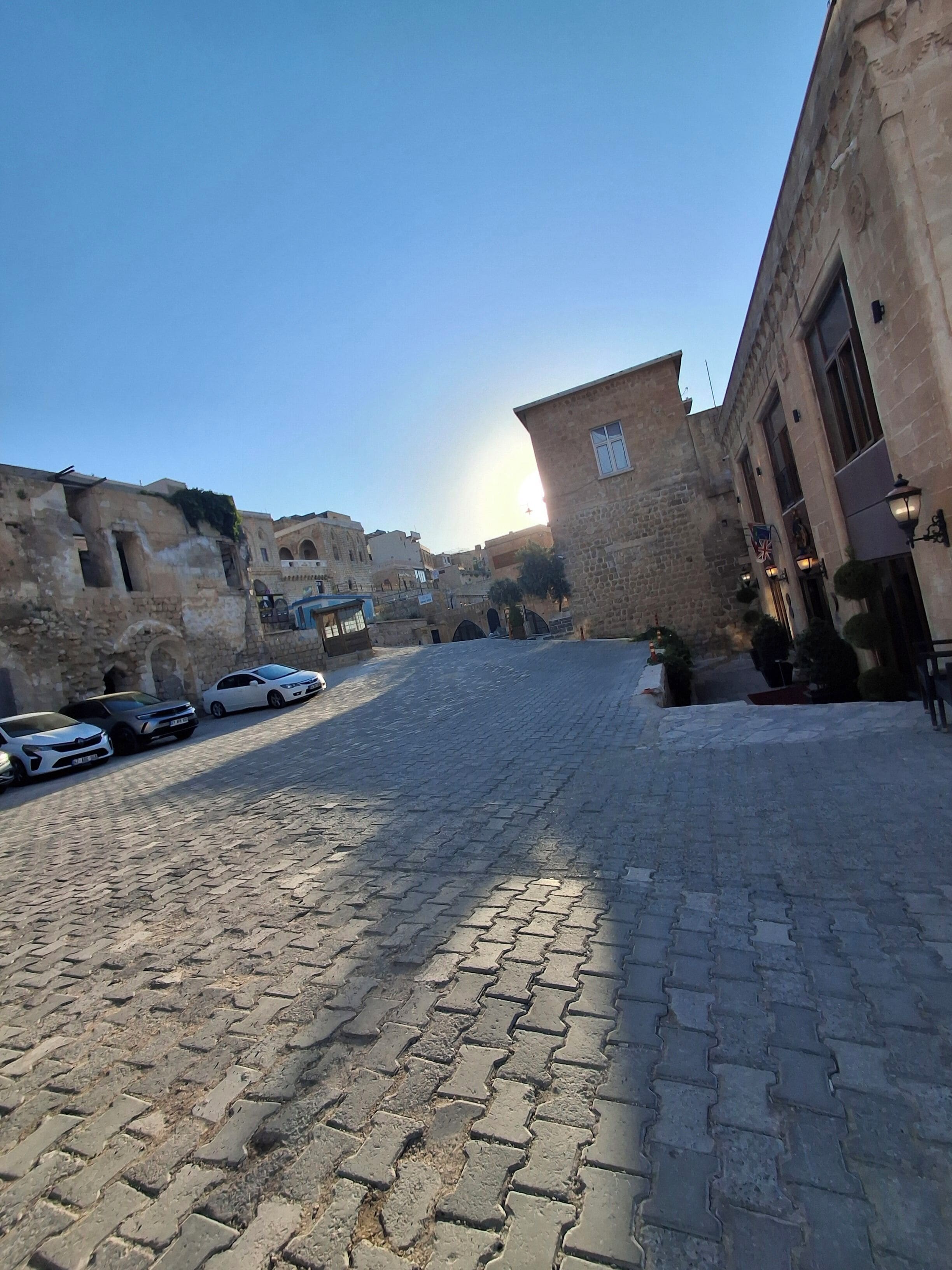 Una calle adoquinada en un pueblo histórico con edificios de piedra, coches aparcados y cielo despejado.
