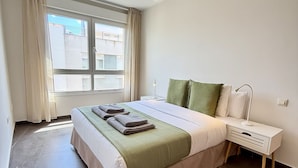 A bedroom with a bed, bedside table, lamp, curtains, and a window. - Dragos Suites 204 (Arrecife)