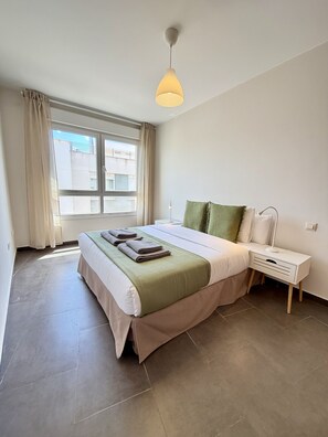 Ein Schlafzimmer mit Bett, Nachttisch, Lampe, Vorhängen und einem Fenster.