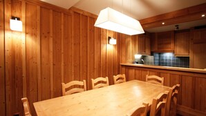 A wooden-paneled room with a large dining table and chairs, a hanging light fixture, and a kitchen area with cabinets. - Spacious Alpine Apartment by the Chairlifts (Les Deux Alpes)