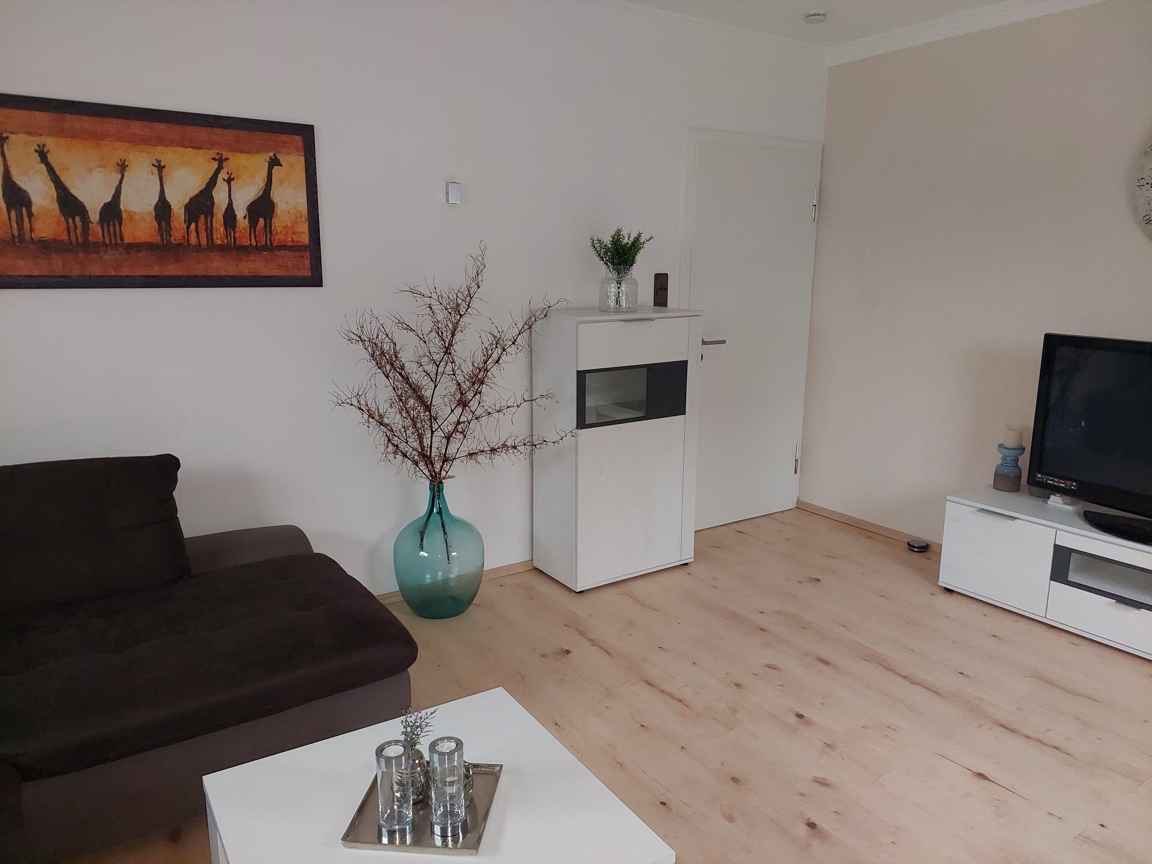Ein Wohnzimmer mit einer Couch, einem Couchtisch mit Salz- und Pfefferstreuern, einem Fernseher auf einem Schrank, einer weißen Kommode, einer Vase mit Ästen und einem Gemälde von Giraffen.