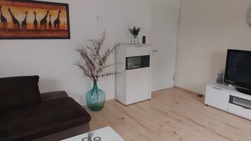 Ein Wohnzimmer mit einer Couch, einem Couchtisch mit Salz- und Pfefferstreuern, einem Fernseher auf einem Schrank, einer weißen Kommode, einer Vase mit Ästen und einem Gemälde von Giraffen.