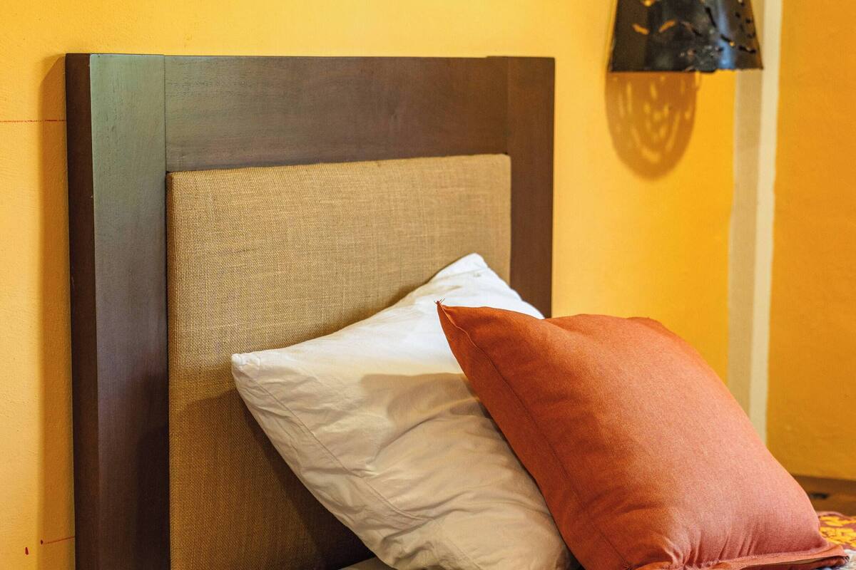 Un lit avec une tête de lit, deux oreillers, une couverture à motifs et une lampe fixée au mur.