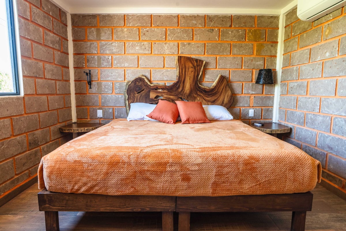 Une chambre avec un mur en briques, une tête de lit en bois, un lit recouvert d’une couverture orange, deux tables de chevet et une petite table de nuit avec une lampe.