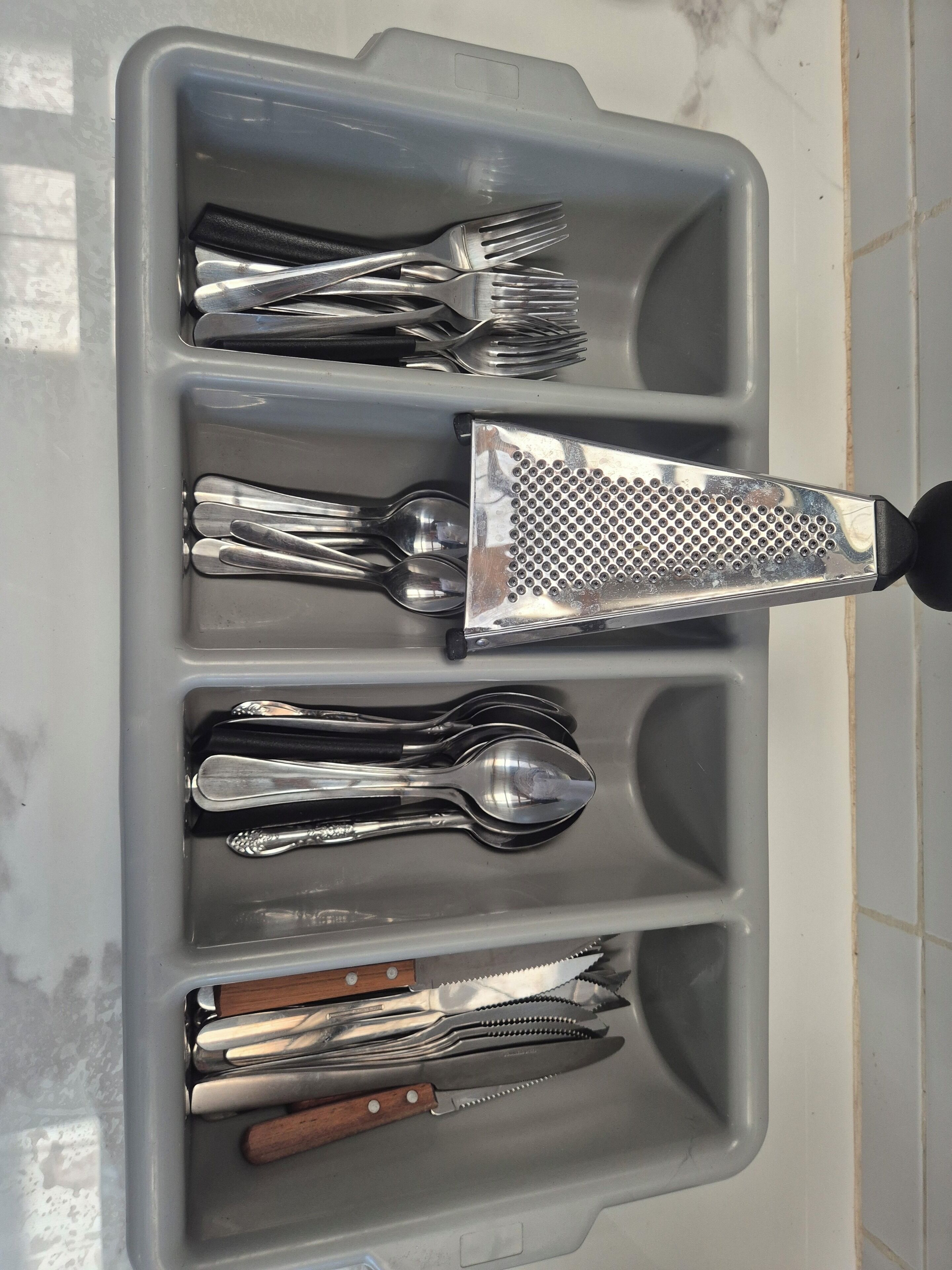 Uma gaveta com diversos utensílios, incluindo garfos, colheres, pinças e um ralador.