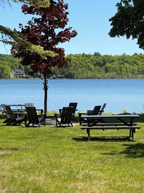 A lakeside view with picnic tables and chairs, a tree, and a building in the background.