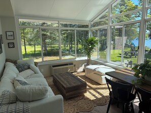 A sunroom with a large window, a white sofa, a wooden coffee table, and a dining table with chairs.
