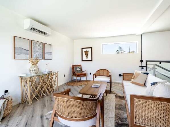 A modern living room with a dining area, wicker furniture, a large window with a view, and a mounted air conditioner.