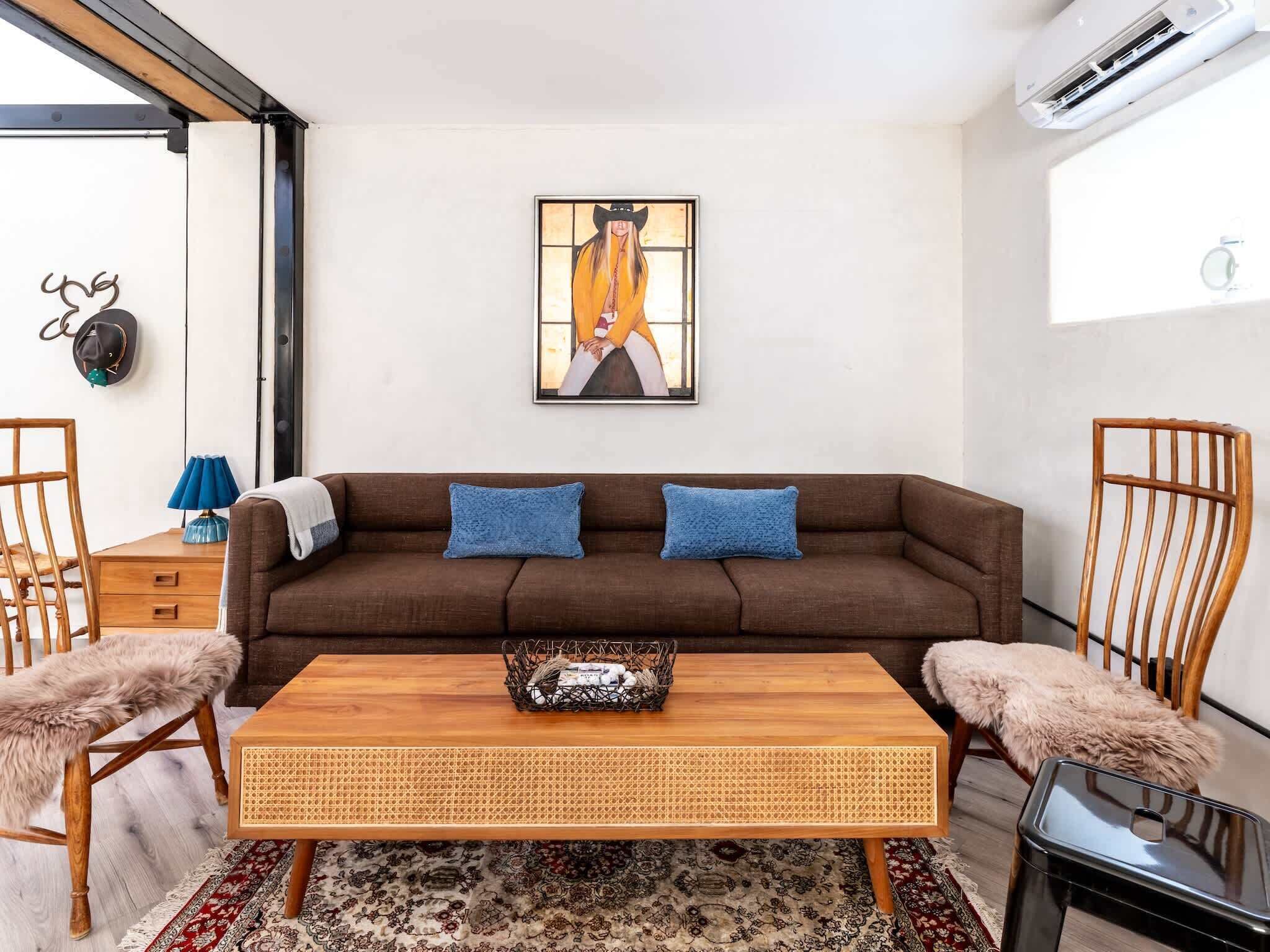 Een moderne woonkamer met een bruine bank, twee blauwe kussens, een houten salontafel, een vloerkleed met een patroon, en een wandairconditioning.