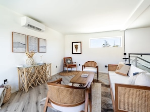 A modern living room with a dining area, wicker furniture, a large window with a view, and a mounted air conditioner.