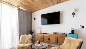 Een woonkamer met een houten plafond, een flatscreen-televisie aan de muur, een houten kast, een koffiezetapparaat en twee bontgekleurde fauteuils rond een glazen salontafel.