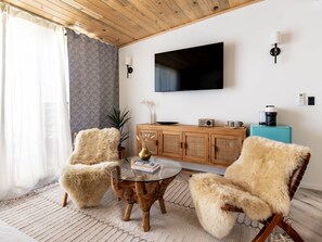 A living room with a wooden ceiling, a flat-screen TV mounted on the wall, a wooden cabinet, a coffee machine, and two fur chairs around a glass coffee table.