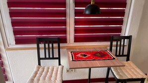Una habitación pequeña con una mesa y dos sillas, un tapete estampado, una lámpara colgante negra y persianas rojas.