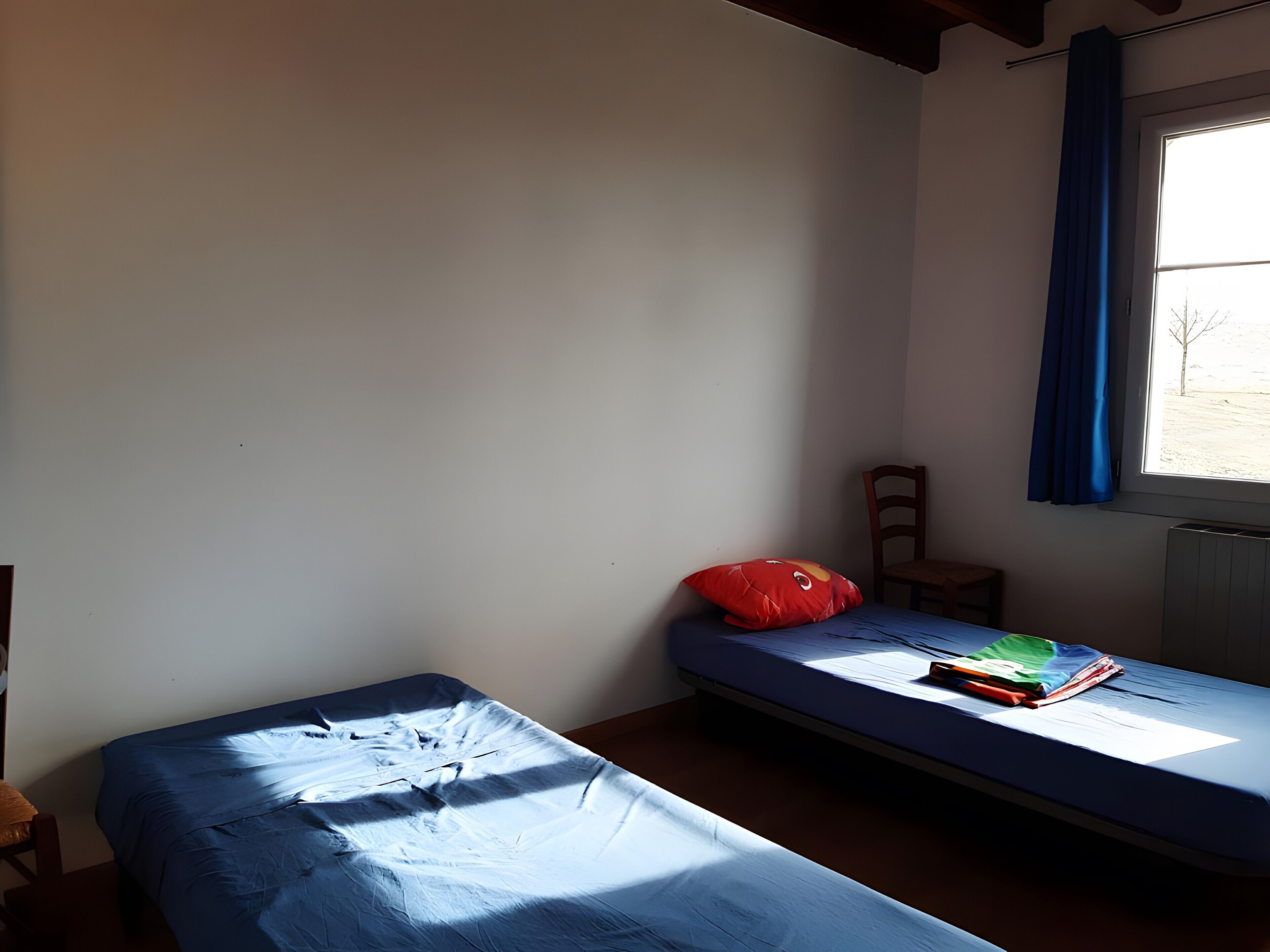 Una habitación pequeña con cama individual, ventana, una silla y almohada roja.