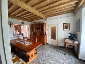 Um quarto com uma mesa de jantar de madeira, um armário com louças, uma porta, uma pequena escrivaninha com abajur e um chão ladrilhado.