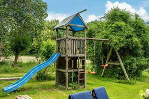 Une structure de jeux en bois, avec un toboggan et des balançoires, installée dans un espace herbeux.