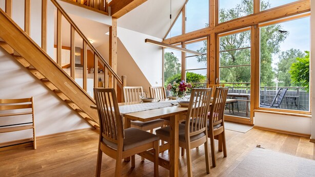 A dining area with a wooden table and chairs, a staircase, and large windows with a view of greenery. - Apartment "Waldkauz" with Private Balcony (Putbus)