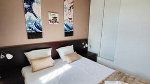 Un lit bien fait, avec du linge de lit blanc et une couverture marron moelleuse, une tête de lit en bois foncé et deux œuvres d’art accrochées au mur représentant des scènes traditionnelles.