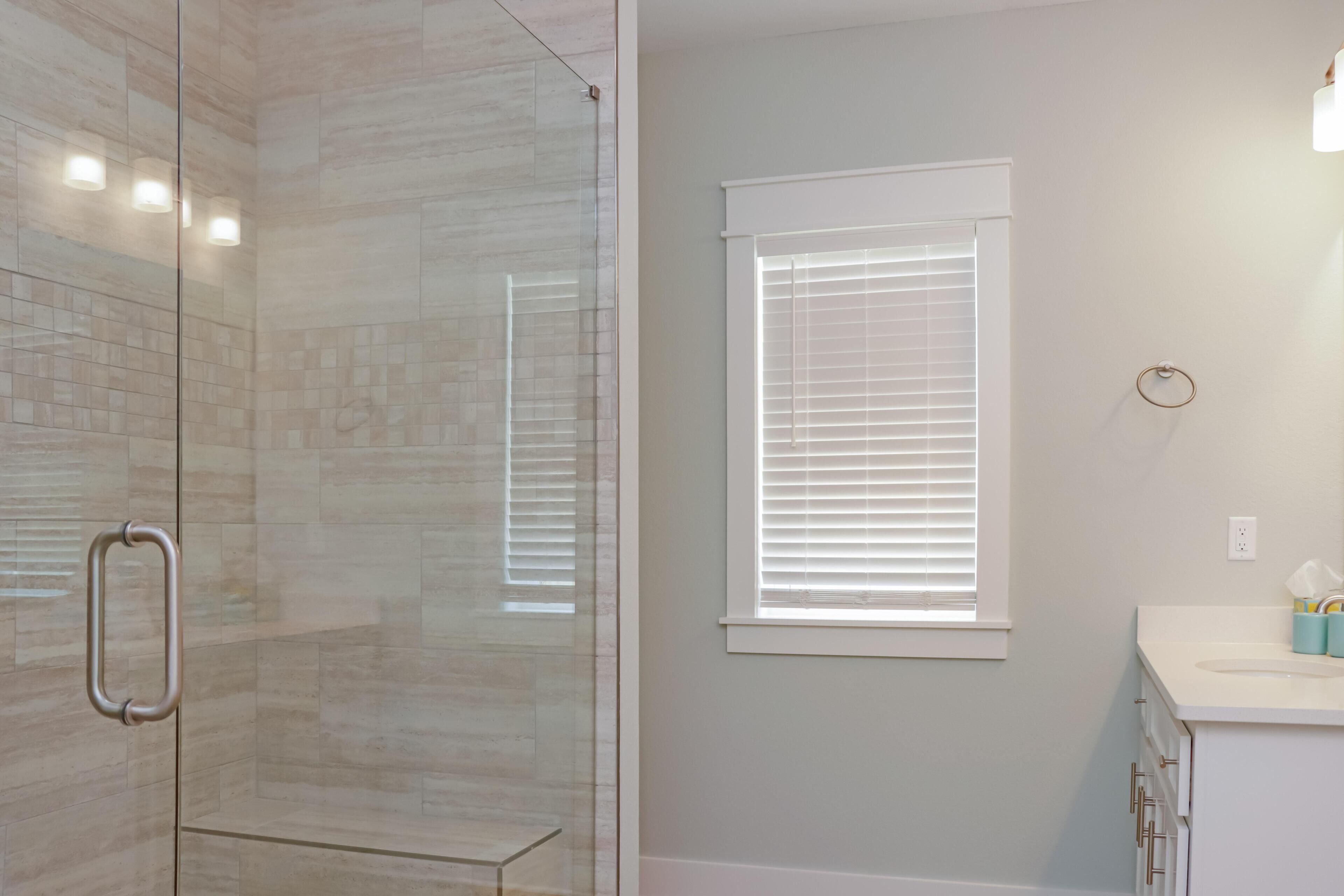A bathroom with a glass shower enclosure, a window with blinds, and a white sink with a mirror.