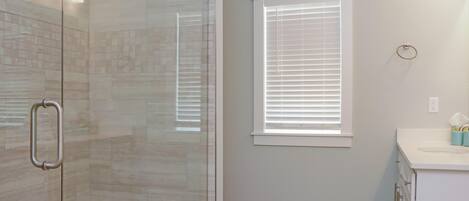 A bathroom with a glass shower enclosure, a window with blinds, and a white sink with a mirror.