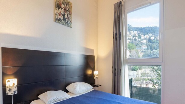 A hotel room with a bed, a nightstand, a lamp, a painting, and a window with a view. - Apartment in Grasse With Swimming Pool (Grasse)