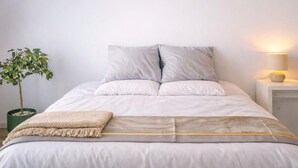 Uma cama bem arrumada com edredom cinza e branco, um pequeno manta bege e uma planta verde em vaso.