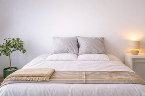 Ein ordentlich bezogenes Bett mit einer grauen und weißen Decke, einer kleinen beigen Tagesdecke und einer Zimmerpflanze im Topf.