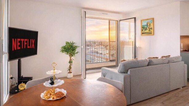 Uma sala de estar moderna com um sofá cinzento, uma TV exibindo 'NETFLIX', uma mesa de jantar com doces, uma planta em vaso e vista para a cidade através de uma porta de vidro.