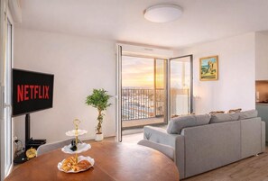 Ein modernes Wohnzimmer mit einer grauen Couch, einem Fernseher, der 'NETFLIX' anzeigt, einem Esstisch mit Gebäck, einer Topfpflanze und Blick auf die Stadt durch eine Glastür.