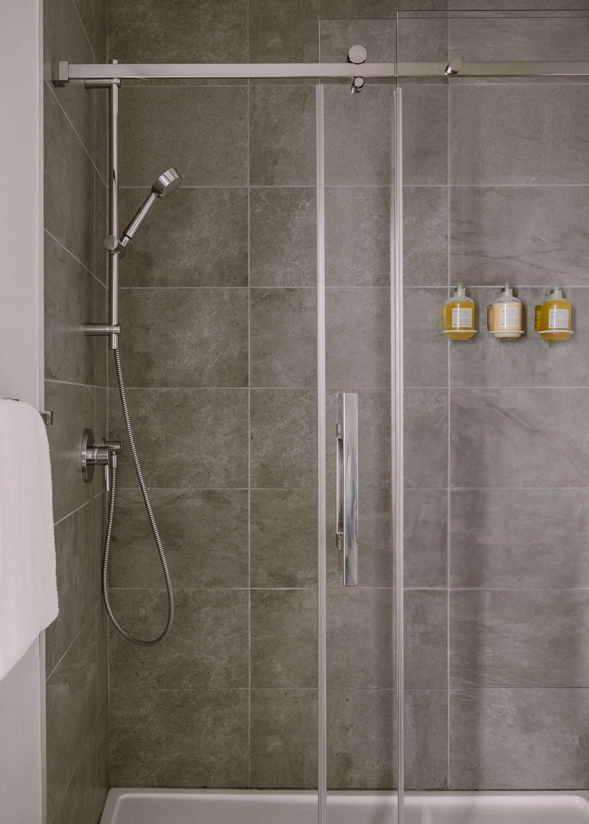 Une douche avec une porte en verre, un pommeau de douche à main et trois bouteilles de shampoing fixées au mur.