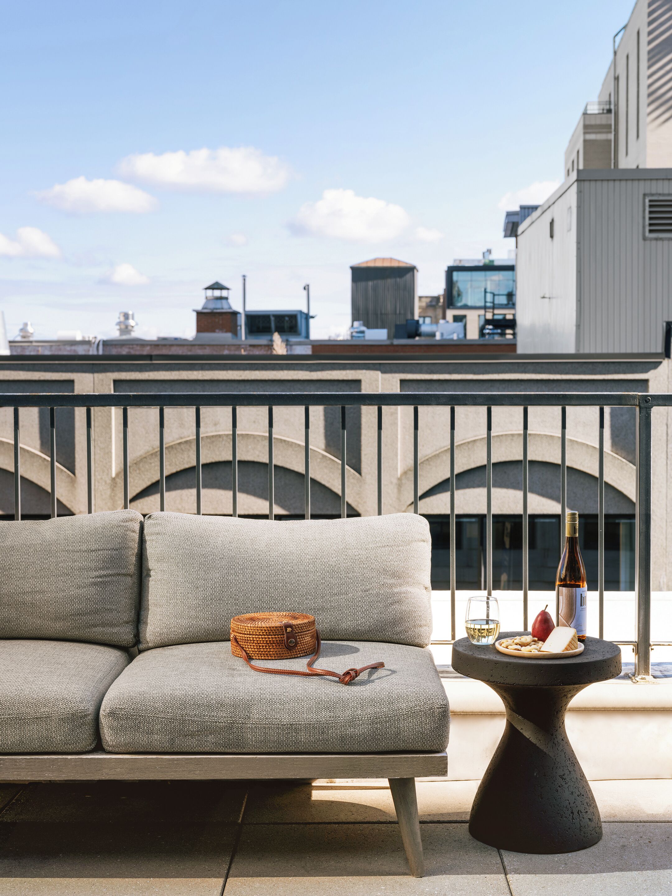 A balcony with a sofa, a small table with a bottle, a glass, and snacks, and a view of buildings in the background.