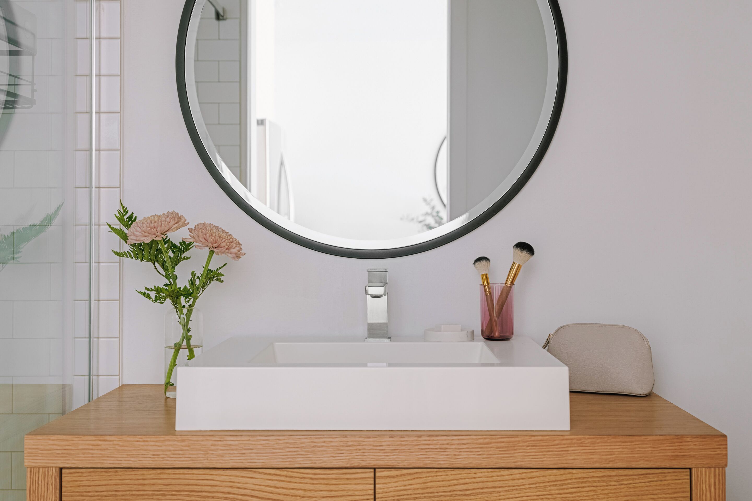 A bathroom with a white sink, a round mirror, a vase with flowers, and a wooden vanity.