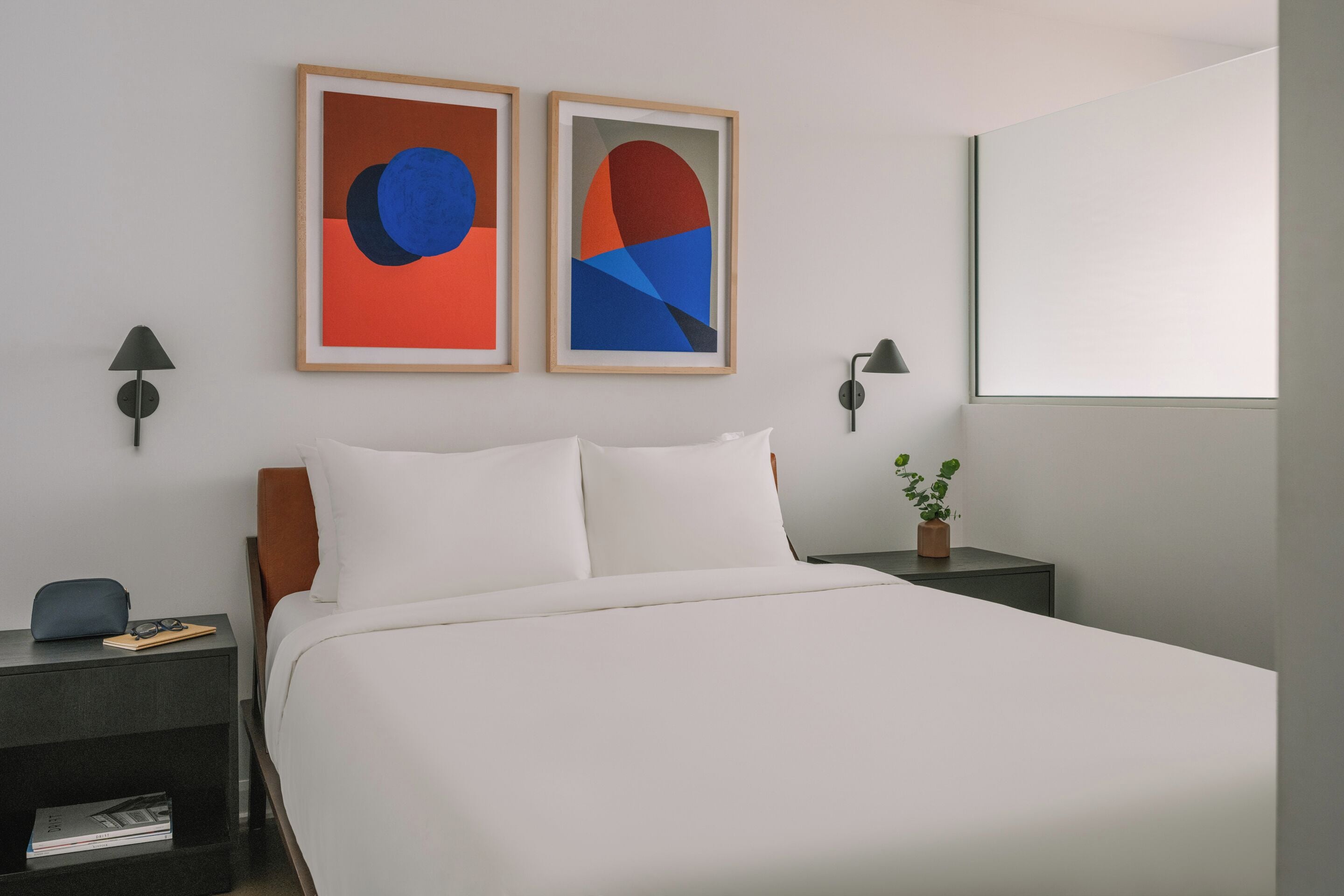Quarto de hotel com uma cama, duas pinturas abstratas emolduradas, uma mesa de cabeceira com abajur, uma pequena mesa com uma planta e uma janela.