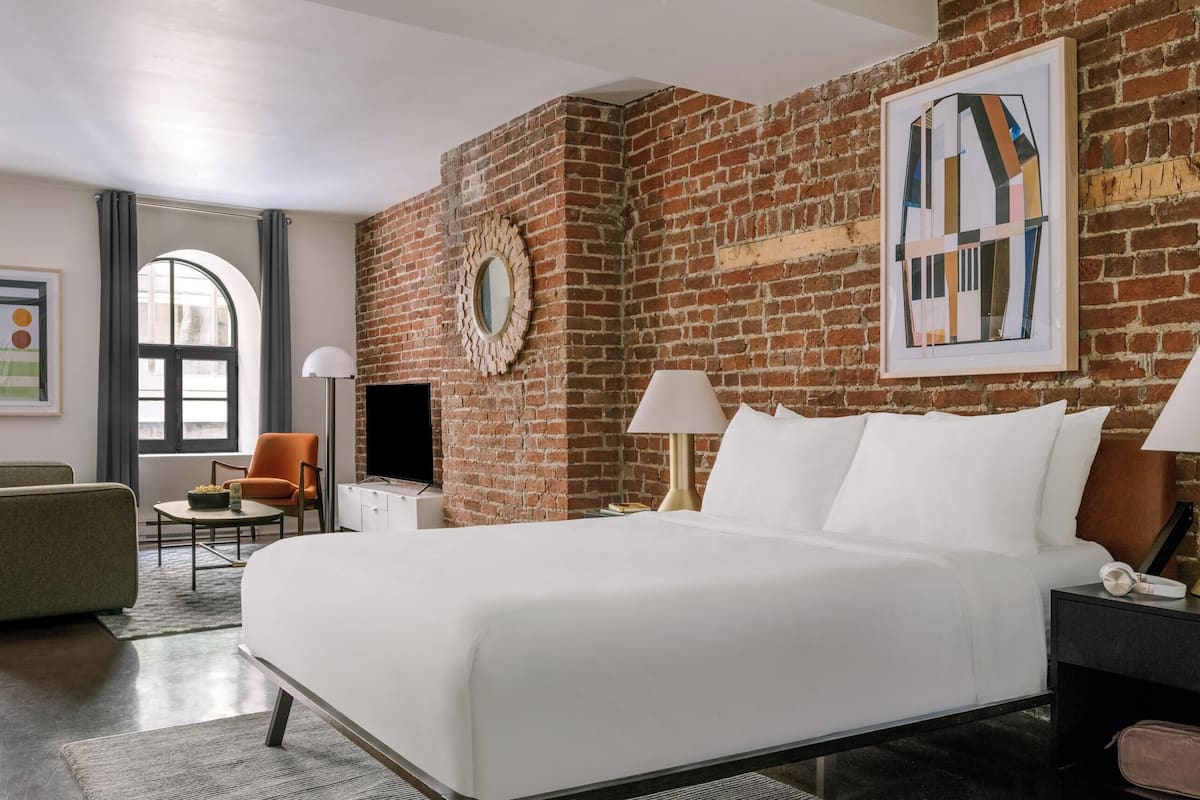 A hotel room with a brick wall, a bed, a TV, a chair, and artwork on the walls.