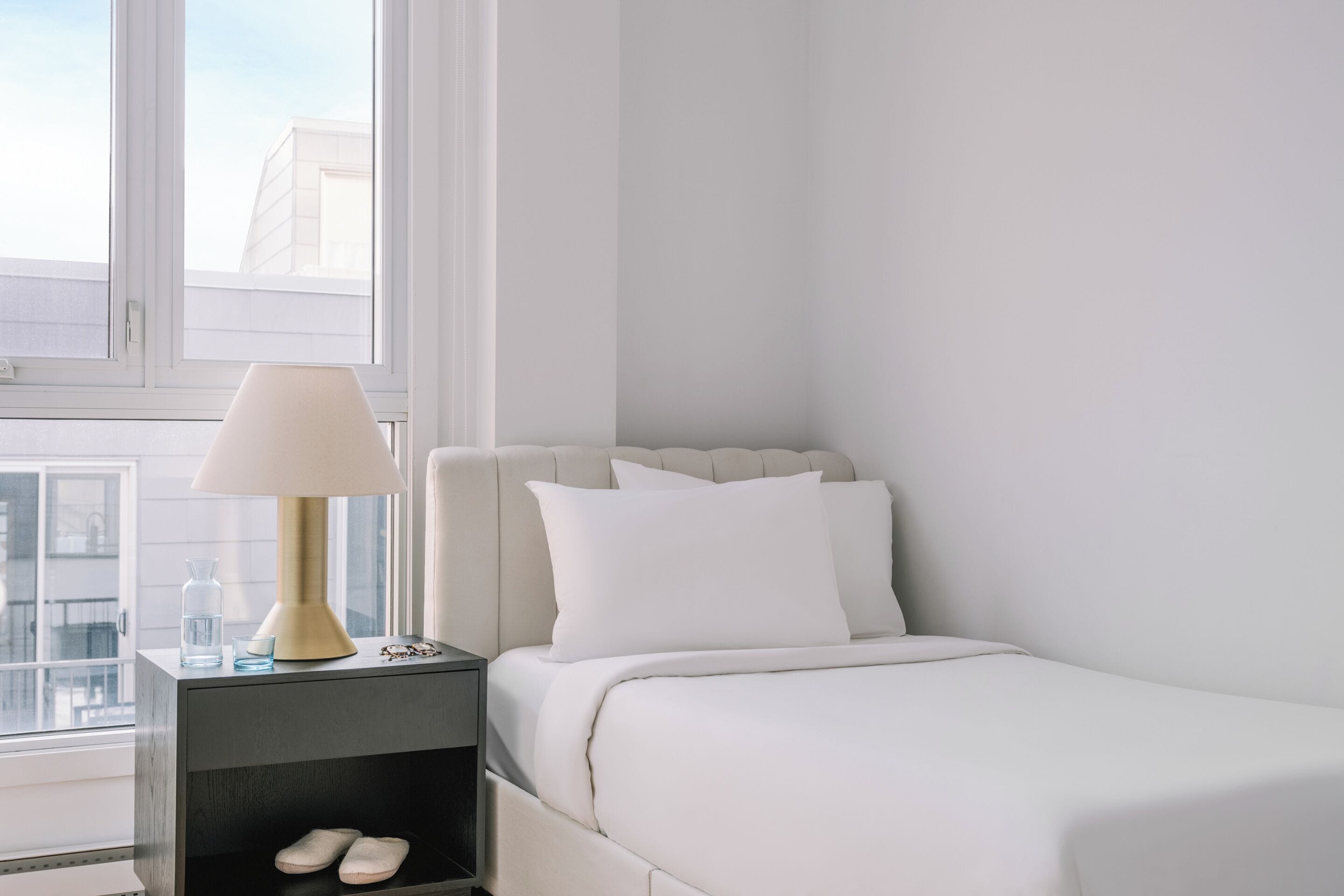 Une chambre d’hôtel avec un lit, une table de chevet avec une lampe, une petite étagère avec des chaussures et une fenêtre donnant sur des immeubles.