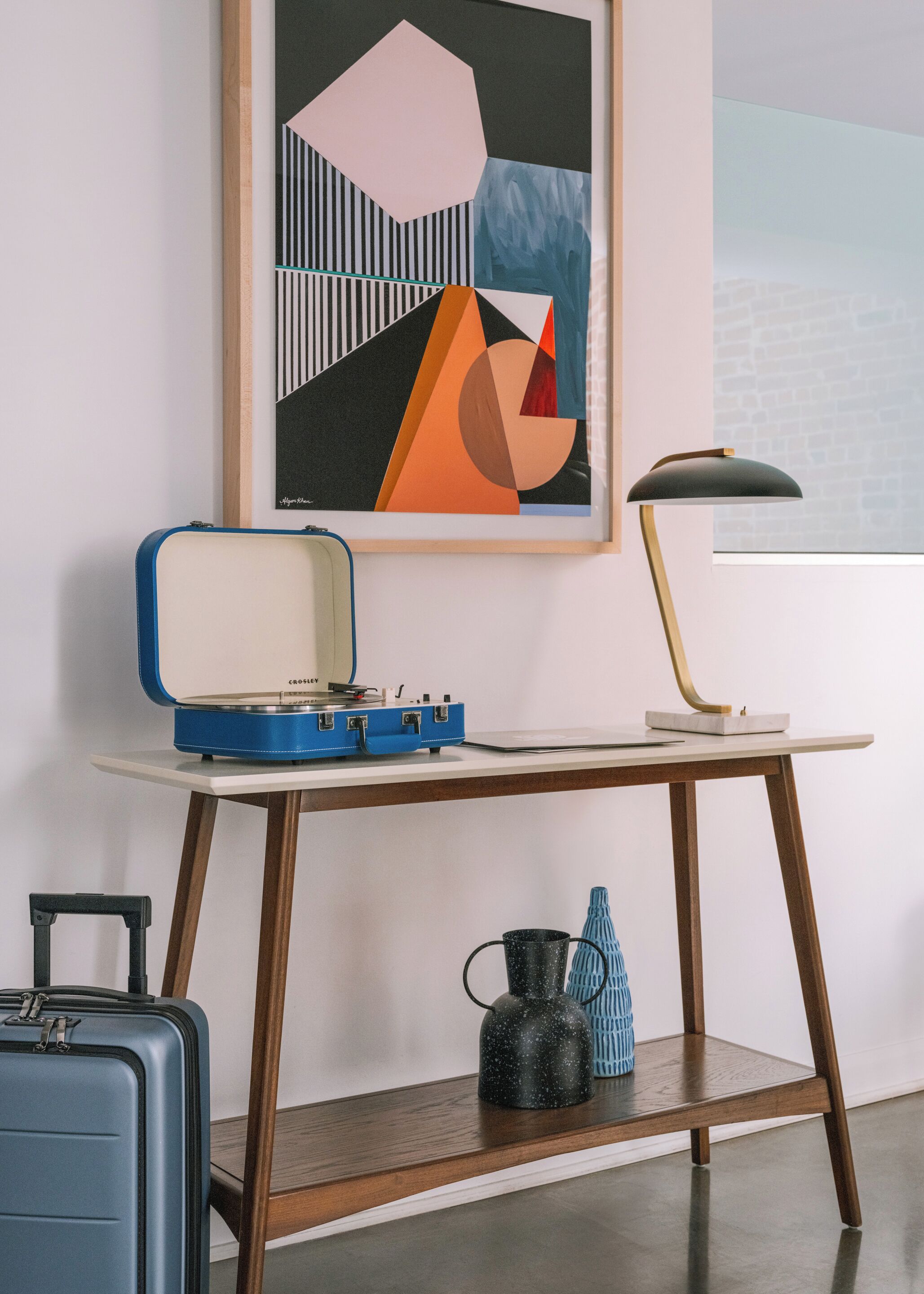 A room with a desk, a blue suitcase, a black kettle, and a framed abstract painting.