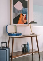 A room with a desk, a blue suitcase, a black kettle, and a framed abstract painting.
