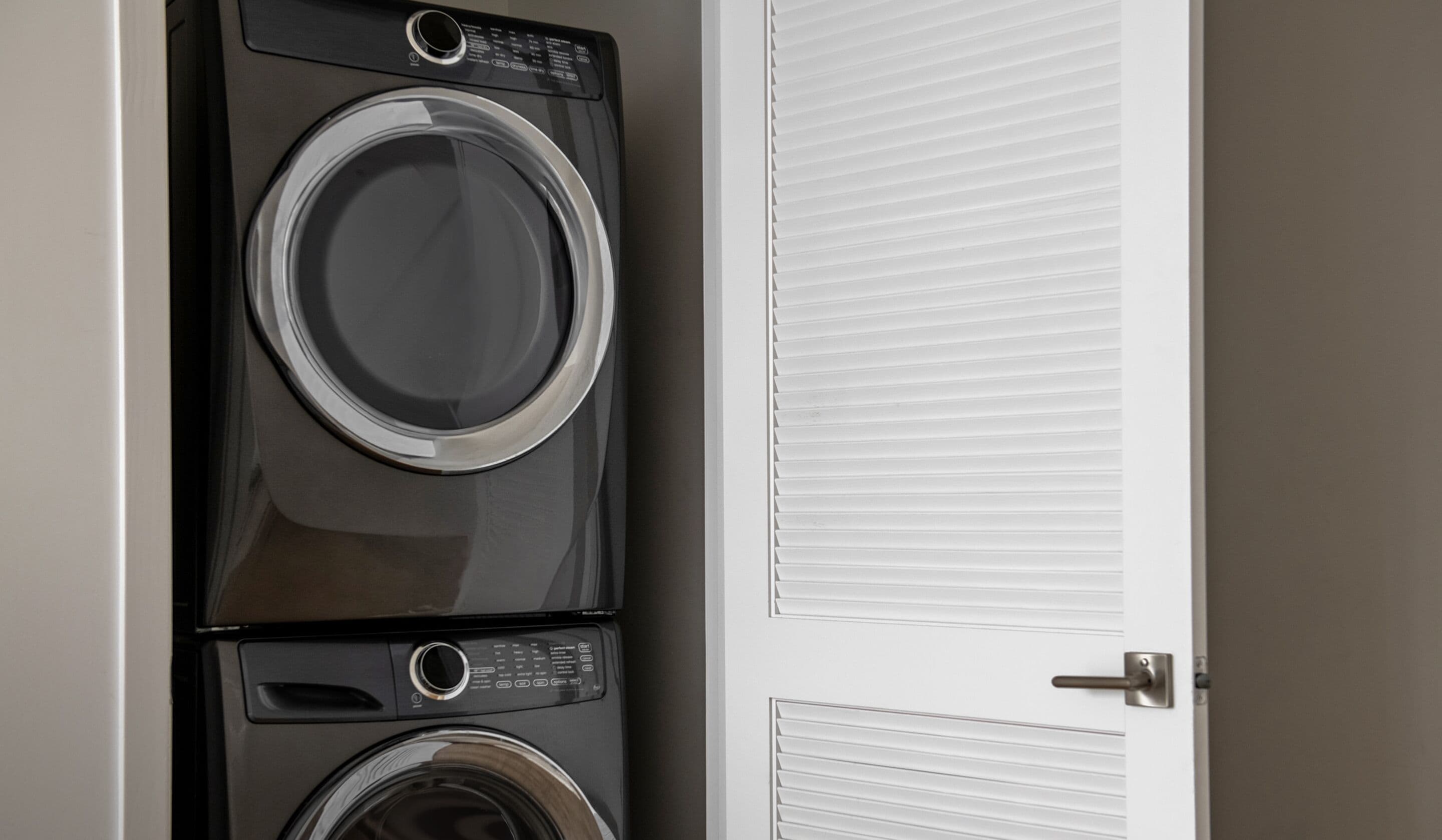 A stacked washer and dryer unit in a room with a closed door and a window with blinds.