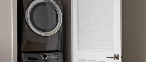 A stacked washer and dryer unit in a room with a closed door and a window with blinds.